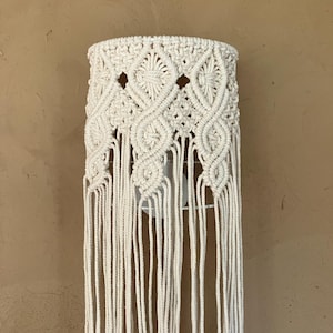 Peut inclure: Un abat-jour en macramé de couleur crème présentant des motifs noués complexes et des découpes en forme de losange. La partie supérieure affiche un tissage dense, tandis que la partie inférieure se compose de longues franges pendantes.
