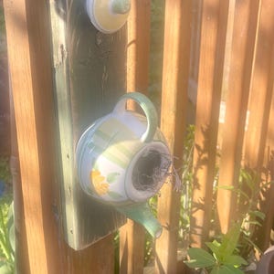 May include: A decorative birdhouse shaped like a teacup, mounted on a wooden post. The birdhouse is white with green stripes and a yellow floral design. A matching lid is attached above. The birdhouse is surrounded by flowers and a wooden fence.