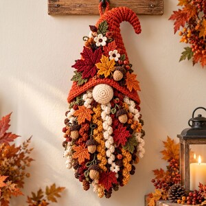 May include: A hanging crochet gnome decoration featuring a tall orange hat adorned with crocheted maple leaves, small white flowers, and acorns. The gnome has a round beige nose and a long, textured beard made of curly yarn in shades of cream, gold, and brown.
