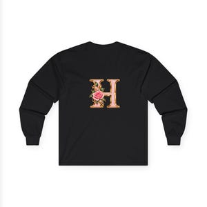 May include: A black long-sleeve shirt featuring a large, metallic gold-toned letter H centered on the chest. The letter is decorated with a detailed illustration of a pink rose and small green leaves attached to the left side of the character.
