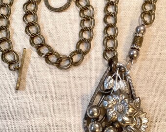 May include: A silver tone pendant necklace with a teardrop-shaped design featuring a flower and berries. The pendant is attached to a brass chain with a large, round, open-link design.