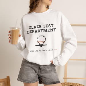 May include: A white crewneck sweatshirt featuring the black text GLAZE TEST DEPARTMENT, a graphic of a pottery vessel on a wheel, and the text RESULTS AFTER FIRING. The sweatshirt is paired with grey denim shorts and a clear cup of iced coffee.