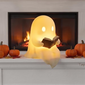 May include: A glowing, off-white ghost-shaped lamp reading a small, dark-colored book. The lamp is positioned on a white mantelpiece alongside several orange pumpkins and autumn leaves. The text TinyTimberToysStore is visible on the mantel.