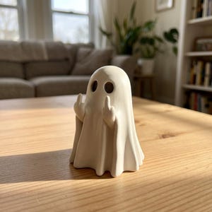 May include: A matte off-white ghost figurine with two dark, circular eye sockets. The ghost has two small arms raised, with each hand extending a middle finger in a gesture. The surface of the figurine appears smooth and solid, standing upright.