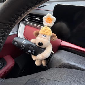 Puede incluir: Un juguete de peluche de perro beige con orejas marrones, que lleva un gorro de ganchillo amarillo rematado con una flor blanca y naranja, está envuelto alrededor de la palanca de intermitentes negra de plástico de una columna de dirección.