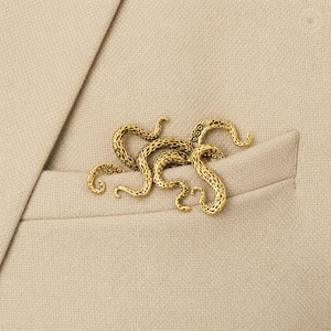 May include: A gold-toned metal brooch shaped like a cluster of octopus tentacles with a textured surface featuring small circular suction cup details. The brooch is pinned to the fabric of a light beige blazer, resting near the chest pocket area.