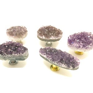 Könnte beinhalten: Fünf amethystfarbene Kristall-Schrankknöpfe mit silbernen und goldenen Metallsockeln. Die Kristalle sind in verschiedenen Violetttönen gehalten und haben eine raue, natürliche Textur.