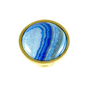 Puede incluir: Una piedra preciosa de ágata redonda, azul y blanca engastada en un marco de metal dorado. El ágata tiene un patrón de remolino de azul y blanco.