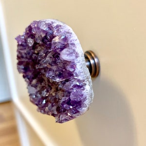 Könnte beinhalten: Ein violetter Amethyst-Kristallknauf mit silberner Basis. Der Knauf ist auf einer weißen Oberfläche montiert.