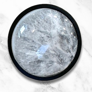 Op de afbeelding: Een ronde, heldere kristal met een zwart metalen frame. De kristal heeft een troebele, witte uitstraling.