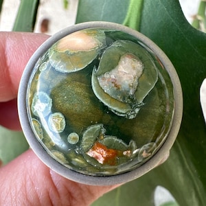 May include: A silver ring with a green, brown, and white moss agate stone. The stone has a unique pattern of swirls and spots.