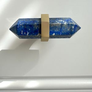 Puede incluir: Un tirador de armario de lapislázuli azul con una banda de metal dorado. La piedra preciosa está facetada y tiene forma rectangular. La ferretería está montada sobre una superficie blanca.