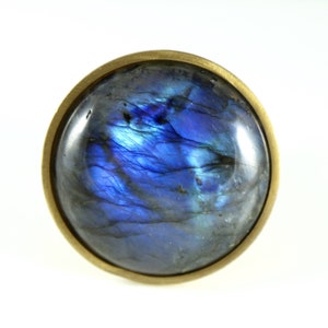 Może przedstawiać: Okrągły, niebieski kamień labradorit osadzony w oprawie z mosiądzu. Kamień ma mieniący się, tęczowy efekt.