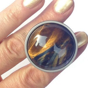 以下が含まれることがあります： 虎目石の大きな丸い石が付いた銀の指輪。石は茶色、金色、黒の模様をしています。