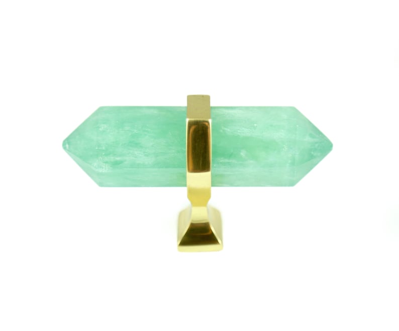 May include: A green crystal cabinet knob with a gold metal base. The crystal is shaped like a long, pointed, double-ended prism.