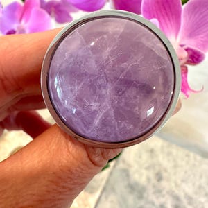 May include: A circular amethyst gemstone, set in a silver-coloured metal bezel. The purple stone displays natural internal patterns. The background features vibrant pink orchid blossoms.