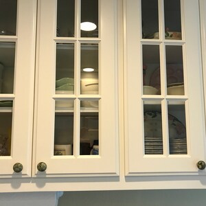 May include: White kitchen cabinets with glass doors and green knobs. The cabinets are filled with dishes and other kitchen items.