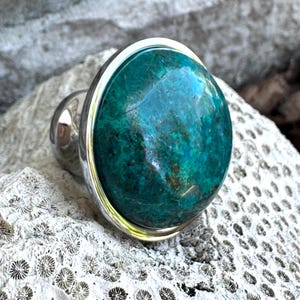 May include: A large, round ring featuring a vibrant teal gemstone set in a silver band. The gemstone has a marbled appearance with shades of green and blue. The ring is displayed on a textured, off-white surface.