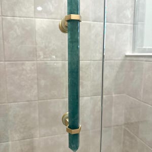 May include: A teal-colored, crystal-shaped shower door handle with gold-colored hexagonal accents. The handle is mounted on a glass shower door, with a tiled wall in the background. The handle is approximately 12 inches long.