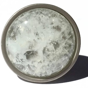 K&ouml;nnte beinhalten: Ein runder, silberfarbener Schrank-Knopf mit einem klaren Glaseinsatz. Das Glas hat eine wei&szlig;e, tr&uuml;be, strukturierte Oberfl&auml;che.