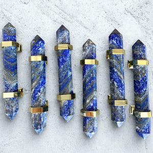 May include: Six blue lapis lazuli crystal wands with gold-colored metal bands. The wands have a pointed tip and a marbled pattern of blue, gray, and white. The gold bands are rectangular and encircle the wands.