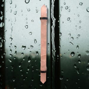 May include: A long, pale pink rose quartz crystal wand with pointed ends, held in place by two black metal bands. The crystal is displayed against a blurred background of a window with raindrops.