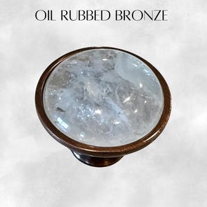 K&ouml;nnte beinhalten: Ein runder, klarer Kristall-Schrankknopf mit einer &ouml;lge&ouml;lten Bronzebasis. Der Knopf hat eine strukturierte Oberfl&auml;che und eine glatte, polierte Oberfl&auml;che.