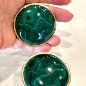 Puede incluir: Dos pomos redondos de malaquita verde con ribete de metal dorado. Los pomos tienen un patrón de espiral.