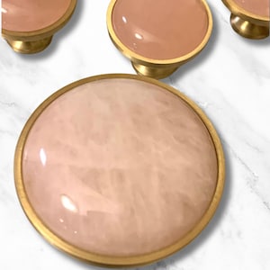 Puede incluir: Un juego de tres pomos redondos para armario, de color rosa y dorado. Los pomos tienen un acabado liso y pulido y una parte superior ligeramente abovedada.