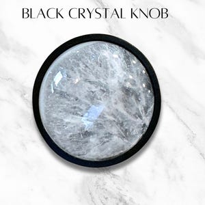 K&ouml;nnte beinhalten: Ein schwarzer Kristallknauf mit einem klaren Kristallkern. Der Knauf ist in einem schwarzen Metallrahmen gefasst.