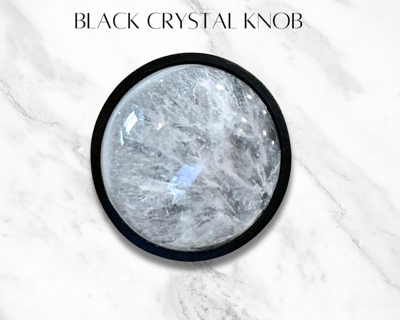 K&ouml;nnte beinhalten: Ein schwarzer Kristallknauf mit einem klaren Kristallkern. Der Knauf ist in einem schwarzen Metallrahmen gefasst.