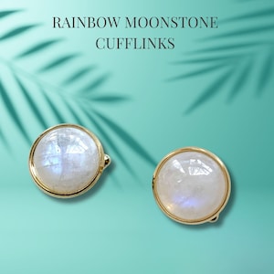 May include: A pair of gold cufflinks with round, iridescent moonstone gemstones. The cufflinks are set against a turquoise background.