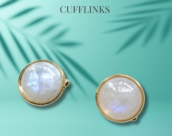 May include: A pair of gold cufflinks with round, iridescent moonstone gemstones. The cufflinks are set against a turquoise background.