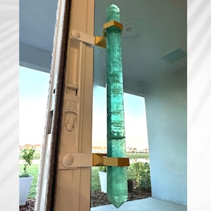Op de afbeelding: Een grote, langwerpige, turquoise kristallen deurklink met goudkleurige metalen beugels. Het kristal is facetgeslepen en heeft een puntige boven- en onderkant. De klink is gemonteerd op een wit deurkozijn.