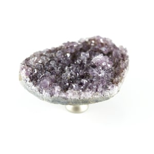 Könnte beinhalten: Eine natürliche Amethyst-Geode, die einen Cluster aus lila Kristallen zeigt. Die Geode ist auf einem kleinen, silberfarbenen Sockel montiert, wodurch ein einzigartiges Dekorationsstück entsteht. Die Kristalle haben ein funkelndes Aussehen, und die Geode ist vor einem weißen Hintergrund platziert.