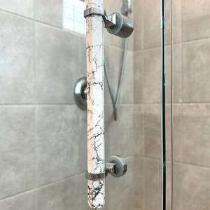 May include: A shower door handle with a white marble design and black veining. The handle is rectangular and attached to the glass door with silver metal brackets. The background shows a shower head and light gray tiles.