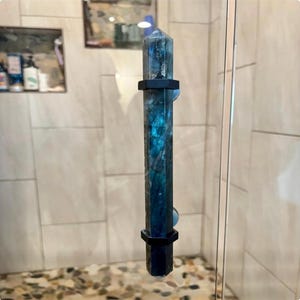 May include: A tall, faceted crystal shower door handle with a blue and green iridescent pattern. The handle is held in place by two black, hexagonal brackets. The handle is mounted on a clear glass shower door.