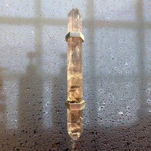 May include: A clear quartz crystal with a light brown hue, featuring two silver-colored metal bands. The crystal is elongated and has a pointed tip. The background is a blurred view of a window with water droplets.