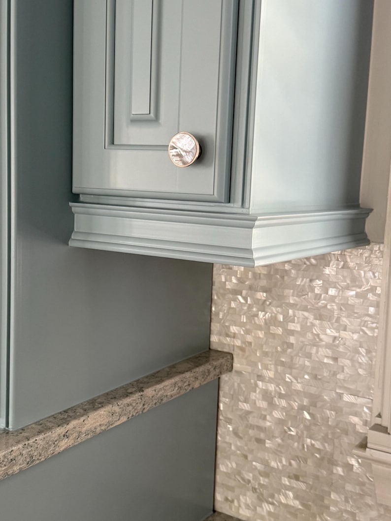 May include: A light blue cabinet door featuring a circular knob with a pearlescent, iridescent finish and a metallic rim. The cabinet has a recessed panel design with a decorative molding at the base, positioned above a speckled countertop.