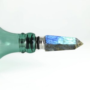 Puede incluir: Una botella de vidrio verde con un tap&oacute;n plateado y negro que presenta un cristal de labradorita azul y gris.