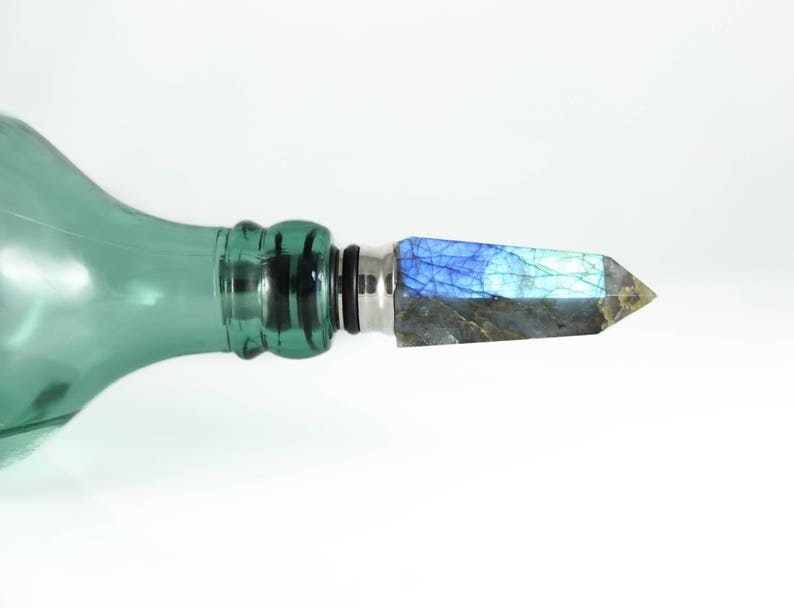 Puede incluir: Una botella de vidrio verde con un tap&oacute;n plateado y negro que presenta un cristal de labradorita azul y gris.