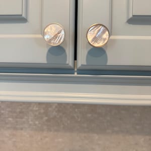 May include: Two circular cabinet knobs featuring a polished mother-of-pearl inlay set within a thin metallic rim. The knobs are mounted on light blue cabinet doors with a recessed panel design, showcasing a smooth, reflective surface texture.