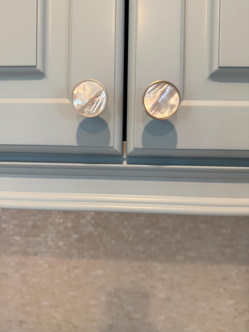 May include: Two circular cabinet knobs featuring a polished mother-of-pearl inlay set within a thin metallic rim. The knobs are mounted on light blue cabinet doors with a recessed panel design, showcasing a smooth, reflective surface texture.