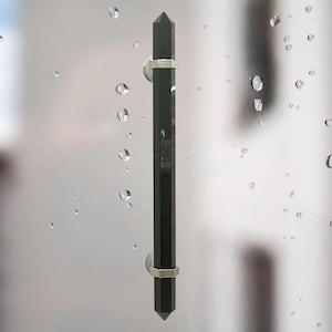 May include: A black, faceted door pull with silver-colored metal accents. The door pull is rectangular with pointed ends and is mounted on a clear surface. Water droplets are visible in the background.