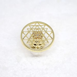 May include: A gold-colored metal pendant featuring a Sri Yantra design. The pendant is circular and has a detailed geometric pattern.