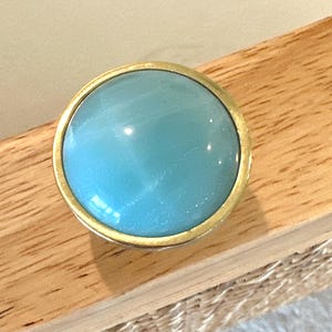 Może przedstawiać: Okrągły, jasnoniebieski kamień kaboszon osadzony w złotej oprawie z metalu. Kamień jest gładki i wypolerowany, odbijający światło. Oprawa znajduje się na drewnianej powierzchni.