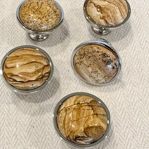 May include: Four round, polished, chrome-plated cabinet knobs with a natural stone inlay. The stone has a brown and beige marbled pattern.