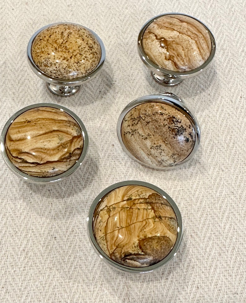 May include: Four round, polished, chrome-plated cabinet knobs with a natural stone inlay. The stone has a brown and beige marbled pattern.