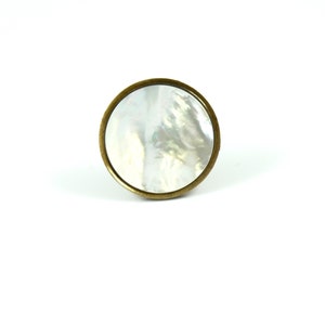 May include: A circular cabinet knob featuring a polished, iridescent white mother-of-pearl inlay set within a smooth, brushed antique brass metal frame. The knob has a simple, flat, round profile with a metallic edge surrounding the shell center.