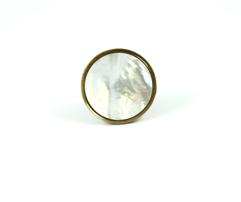 May include: A circular cabinet knob featuring a polished, iridescent white mother-of-pearl inlay set within a smooth, brushed antique brass metal frame. The knob has a simple, flat, round profile with a metallic edge surrounding the shell center.
